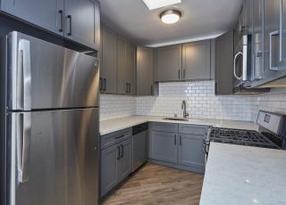 Stainless steel refrigerator next to a wide kitchen countertop with grey cabinets and a cooktop oven