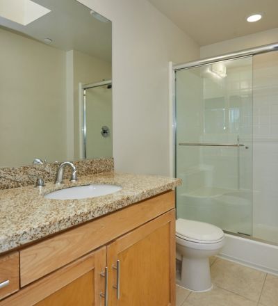 Wall mirror next to a wide marble-finished bathroom countertop with cabinets beside the toilet and glass-enclosed stall