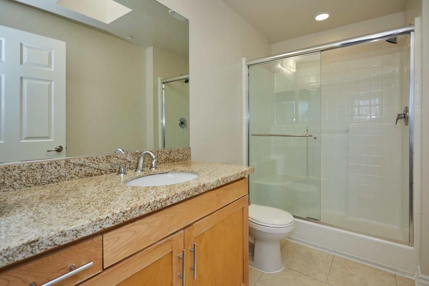 Wall mirror next to a wide marble-finished bathroom countertop with cabinets beside the toilet and glass-enclosed stall